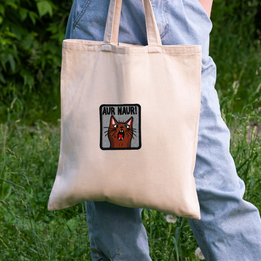 Person holding a beige tote bag with a cat graphic and text in an outdoor setting