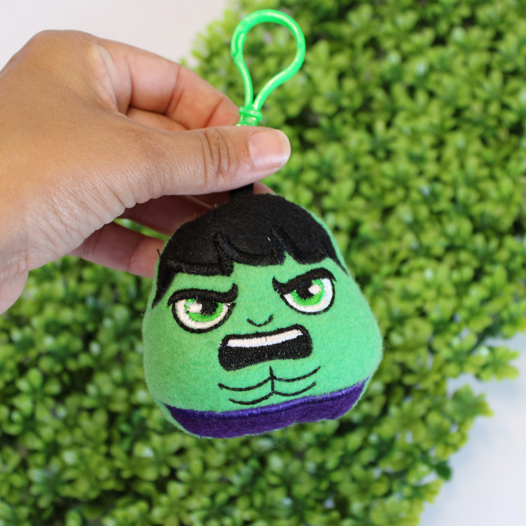 Plush keychain of a green superhero character with a black and purple design, held against a green leafy background.