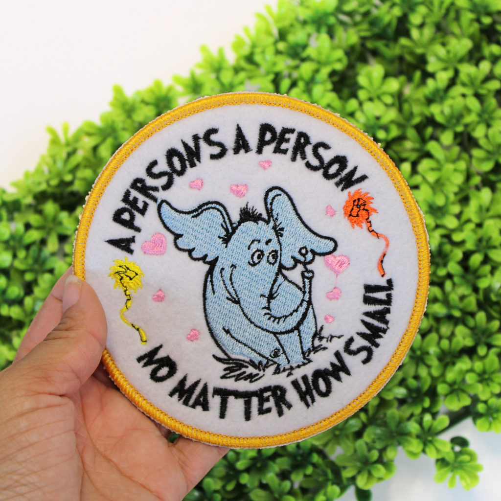 Embroidered patch with a cartoon character and text held in front of green foliage