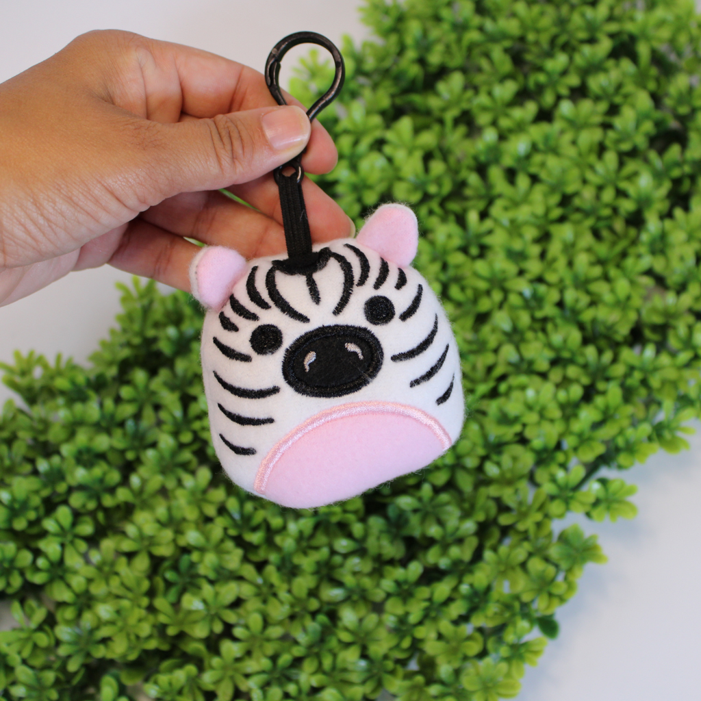 Hand holding a zebra-shaped keychain with pink ears against a green leafy background