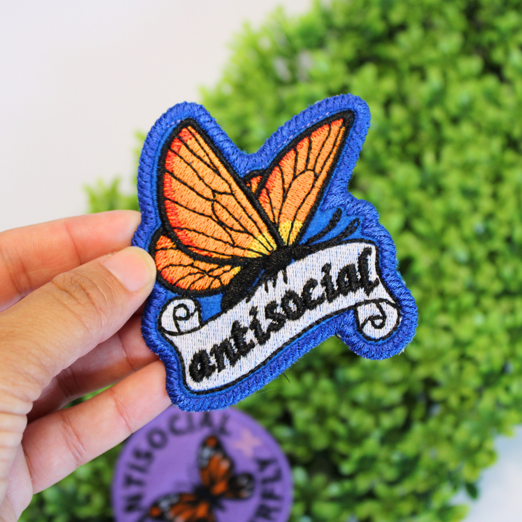 Butterfly patch with 'antisocial' text held in front of green foliage