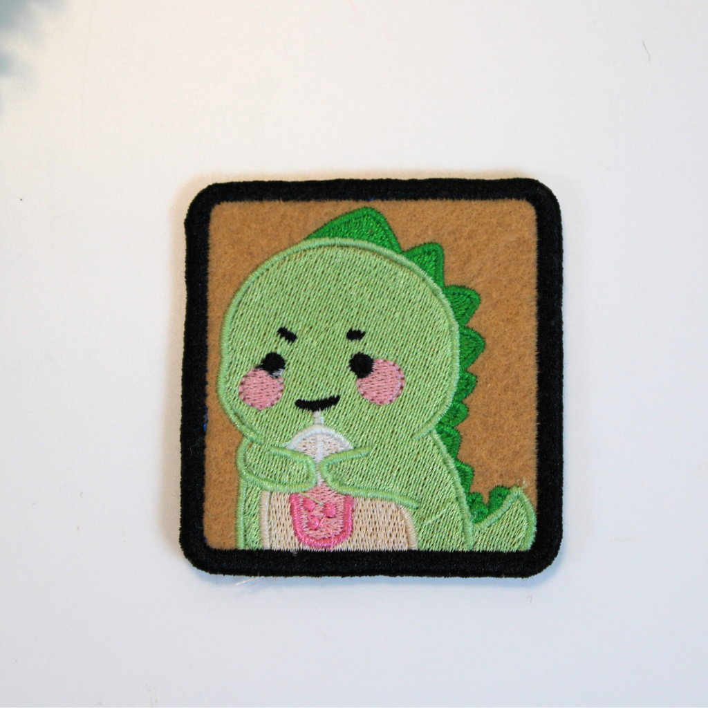 Embroidered patch of a green dinosaur character on a brown square background