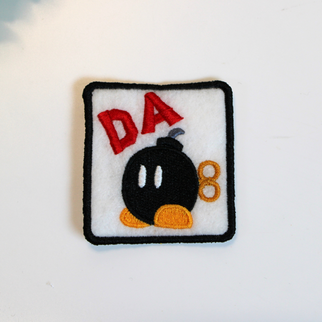 Embroidered patch with a black bomb-like character and 'DA' text on a white background