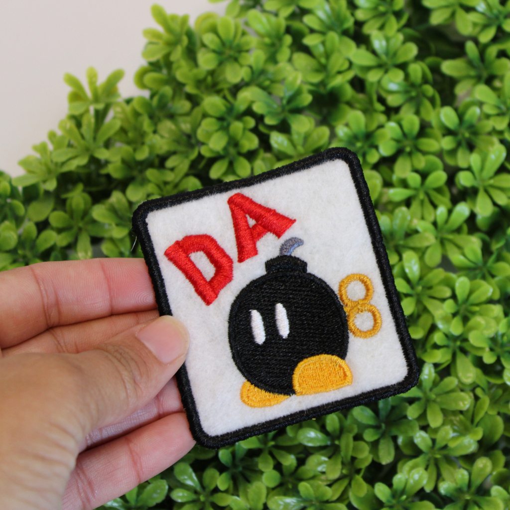 Embroidered patch with a bomb design and 'DA' text held over green foliage