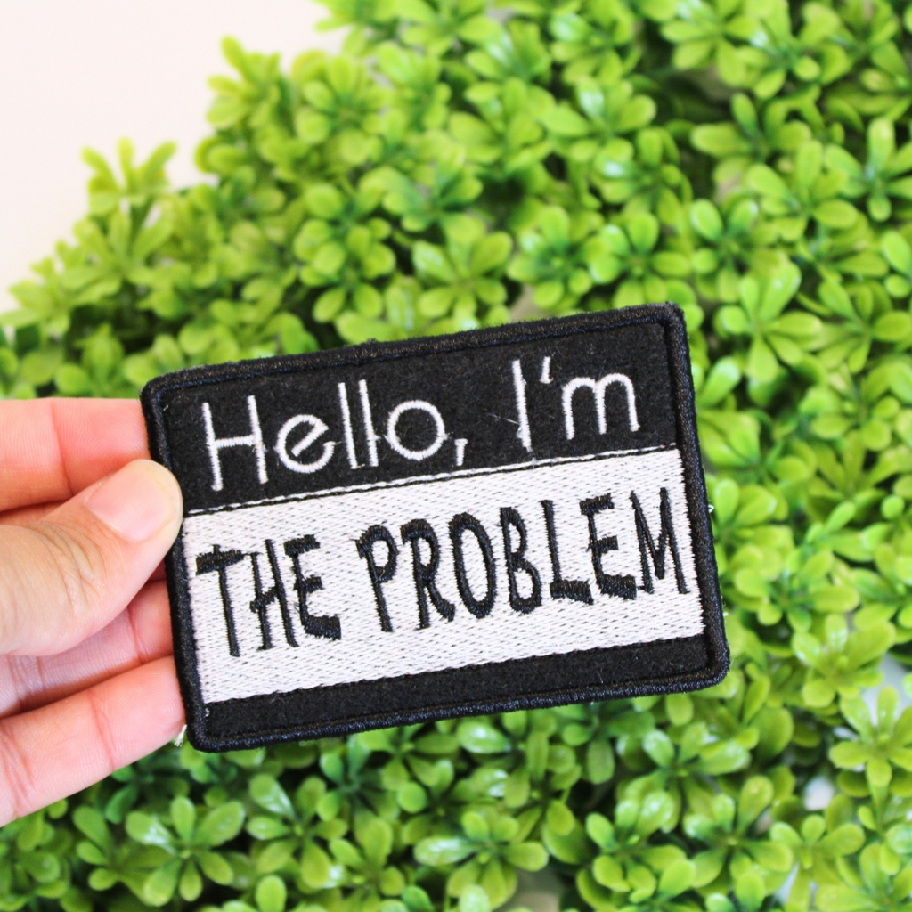 Patch with 'Hello, I'm THE PROBLEM' text held over green foliage