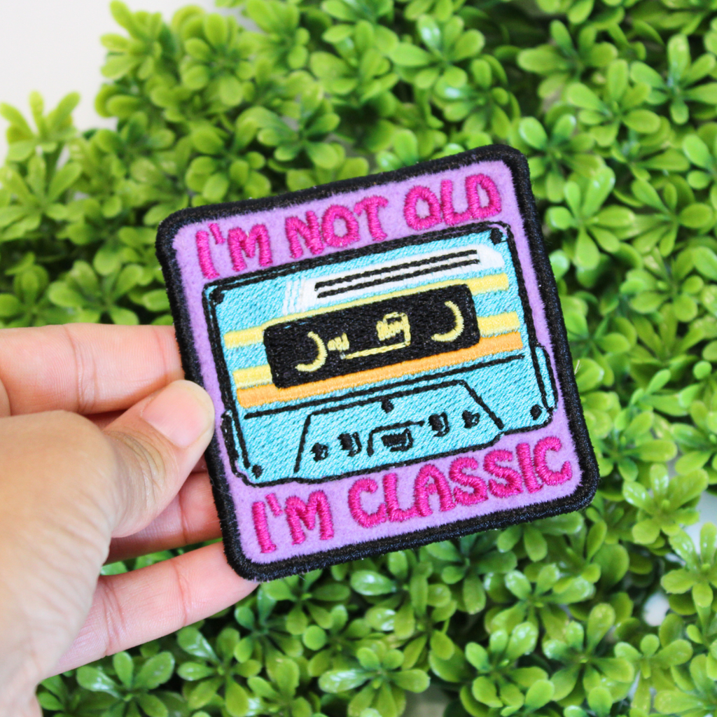 Patch with a cassette tape design and text 'I'm not old, I'm classic' held over green foliage.