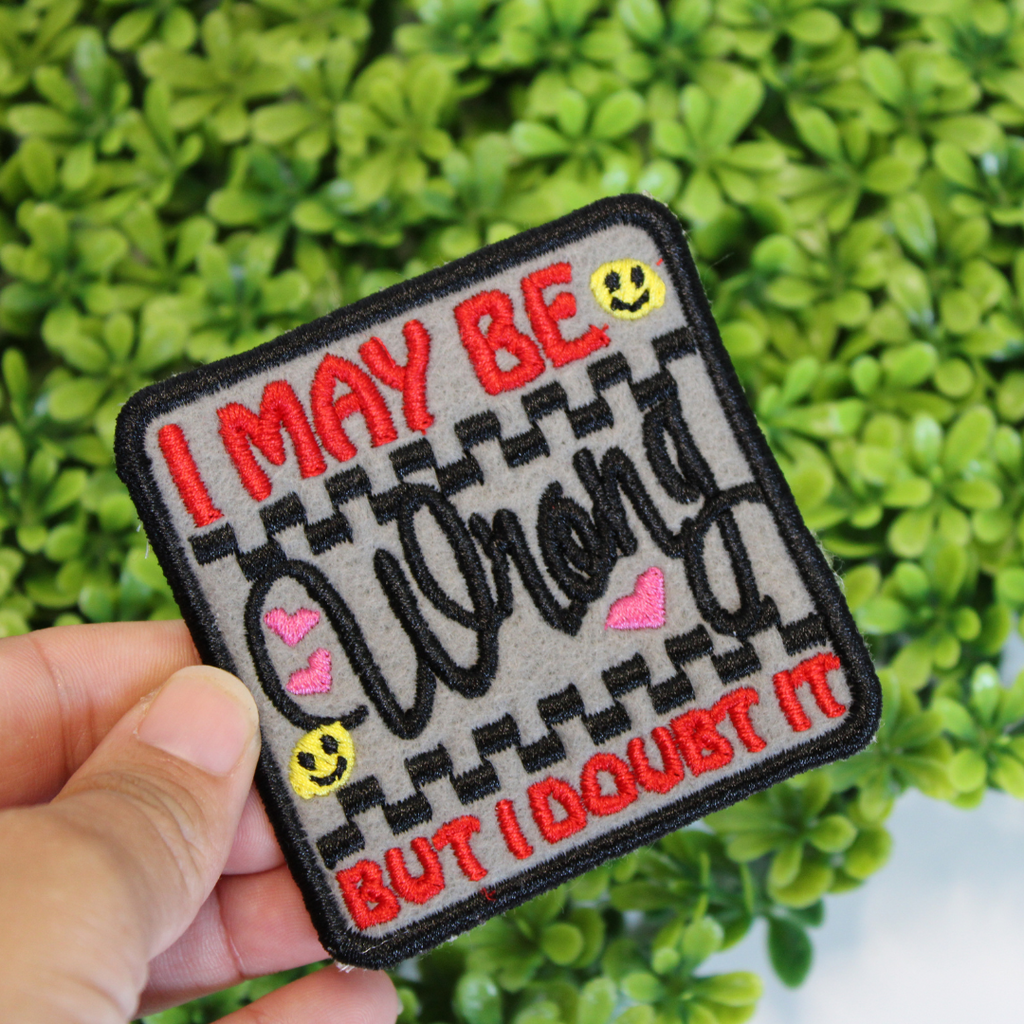 Embroidered patch with text and emojis held in front of green foliage
