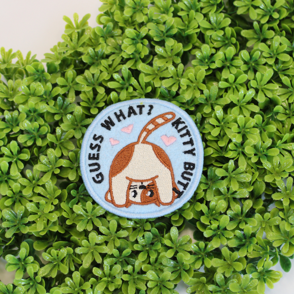 Embroidered patch with a cat design and text 'Guess What? Kitty Butt' on a green leaf background